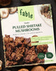 A package of fable brand Pulled Shiitake Mushrooms lies flat on a dark textured surface, surrounded by whole fresh and dried mushrooms in a styled flat-lay arrangement.